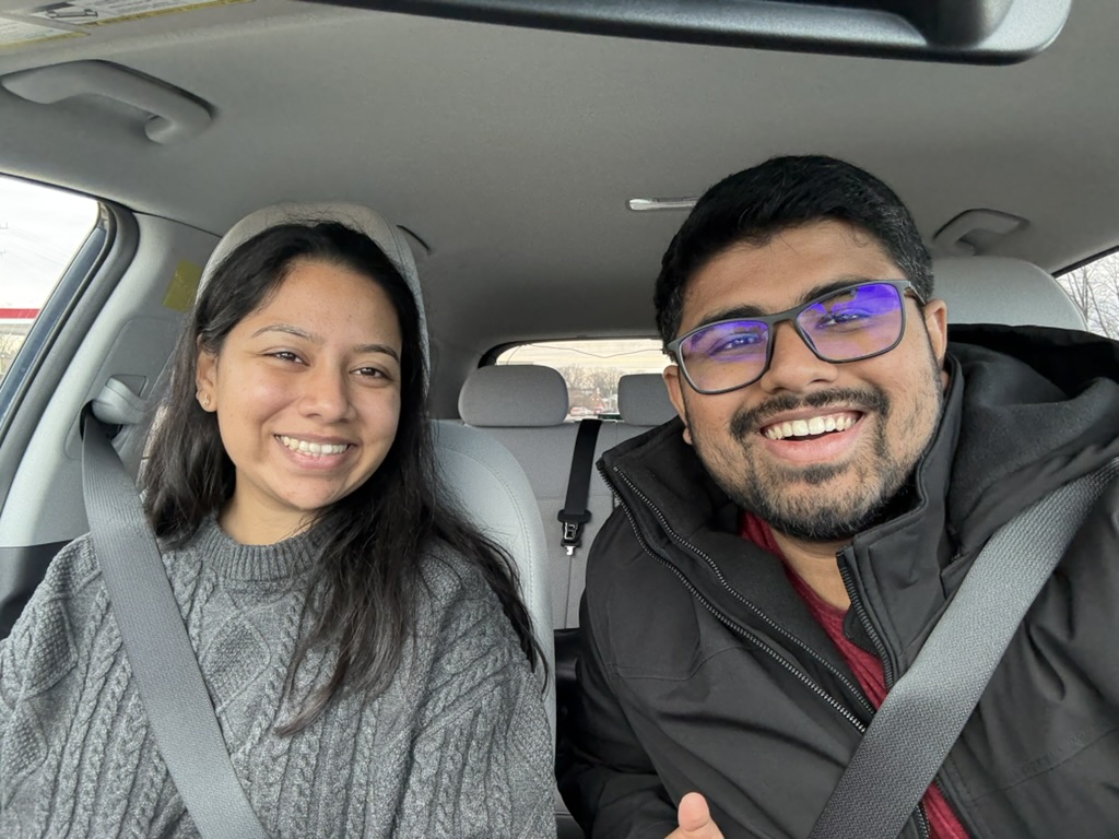 Dakshinya and Karthik smiling together in the car, bundled up in winter coats with their seatbelts on