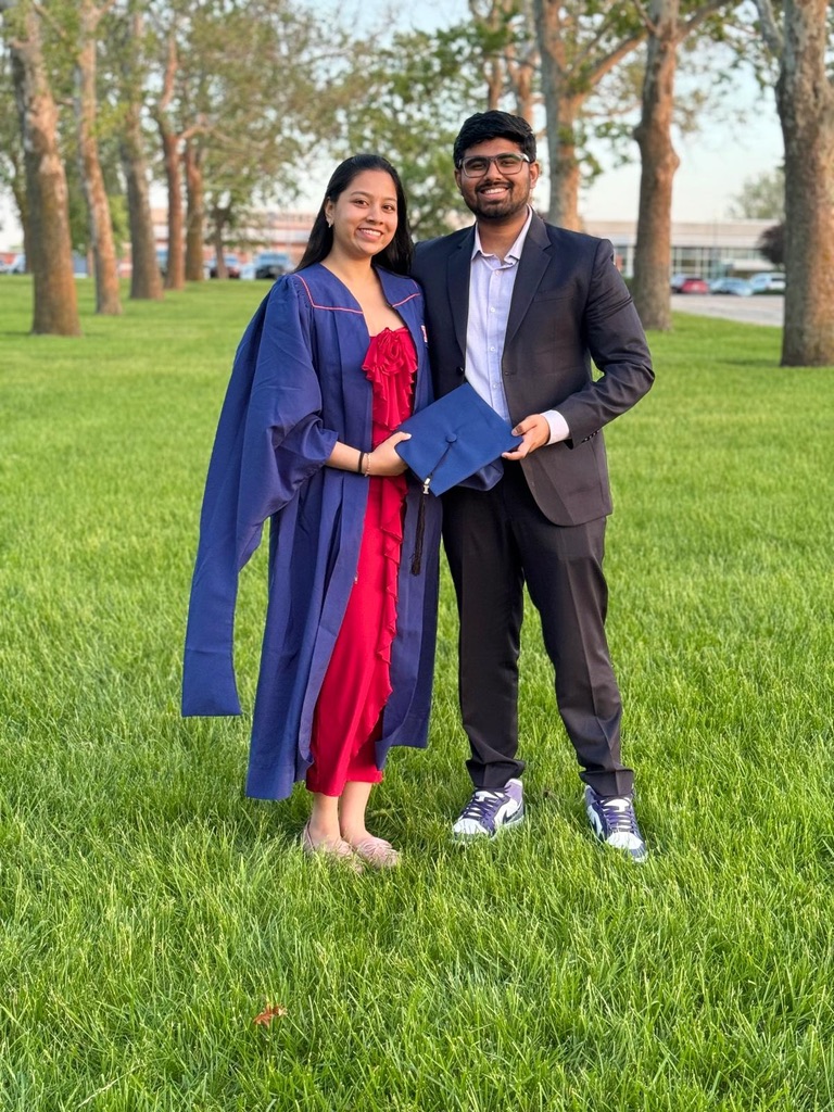 Dakshinya in a blue graduation gown over a red dress, standing on the grass with Karthik in a suit as they hold her cap together