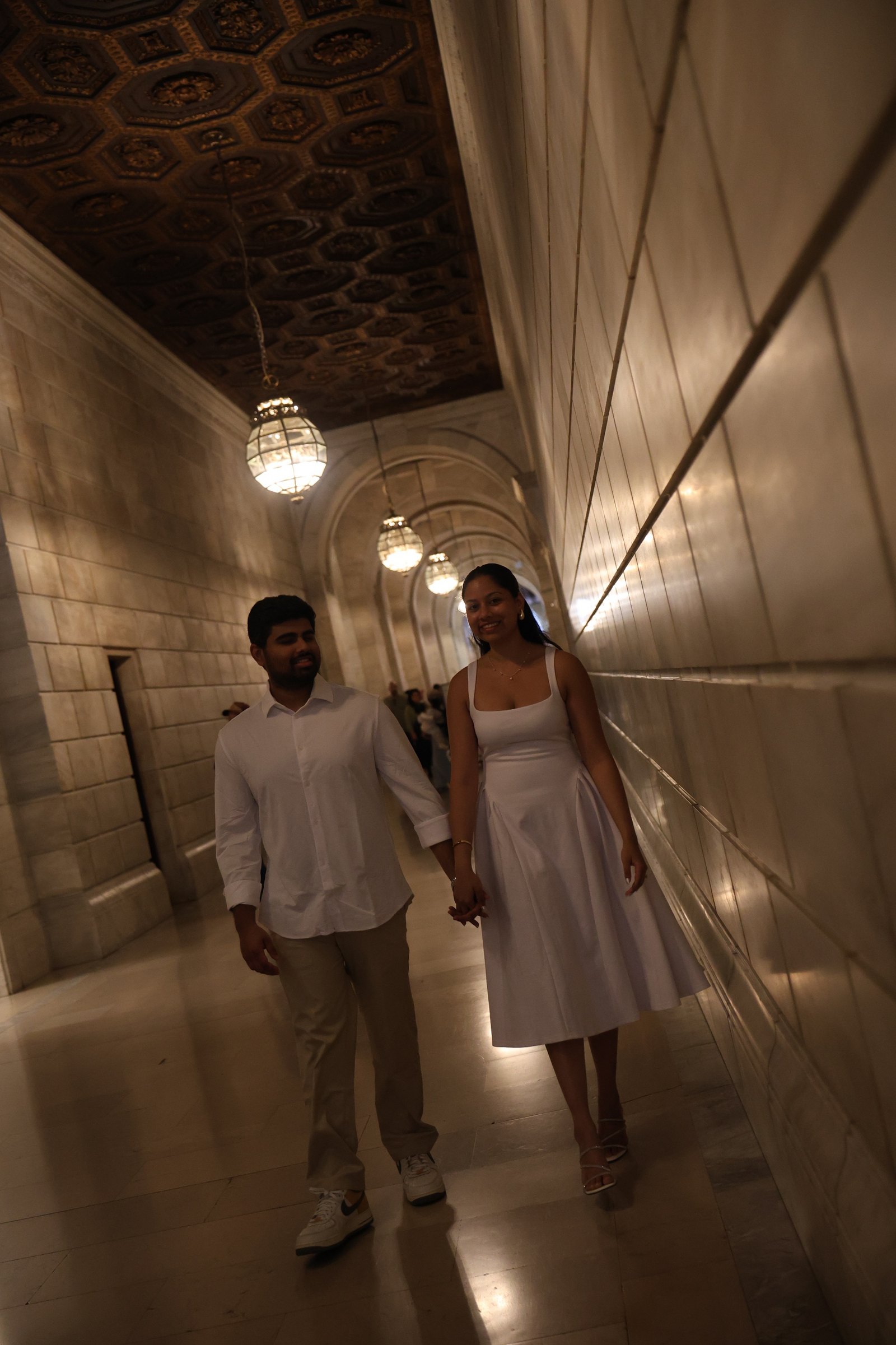 Dakshinya and Karthik walking hand in hand beneath glowing lanterns in a marble hallway