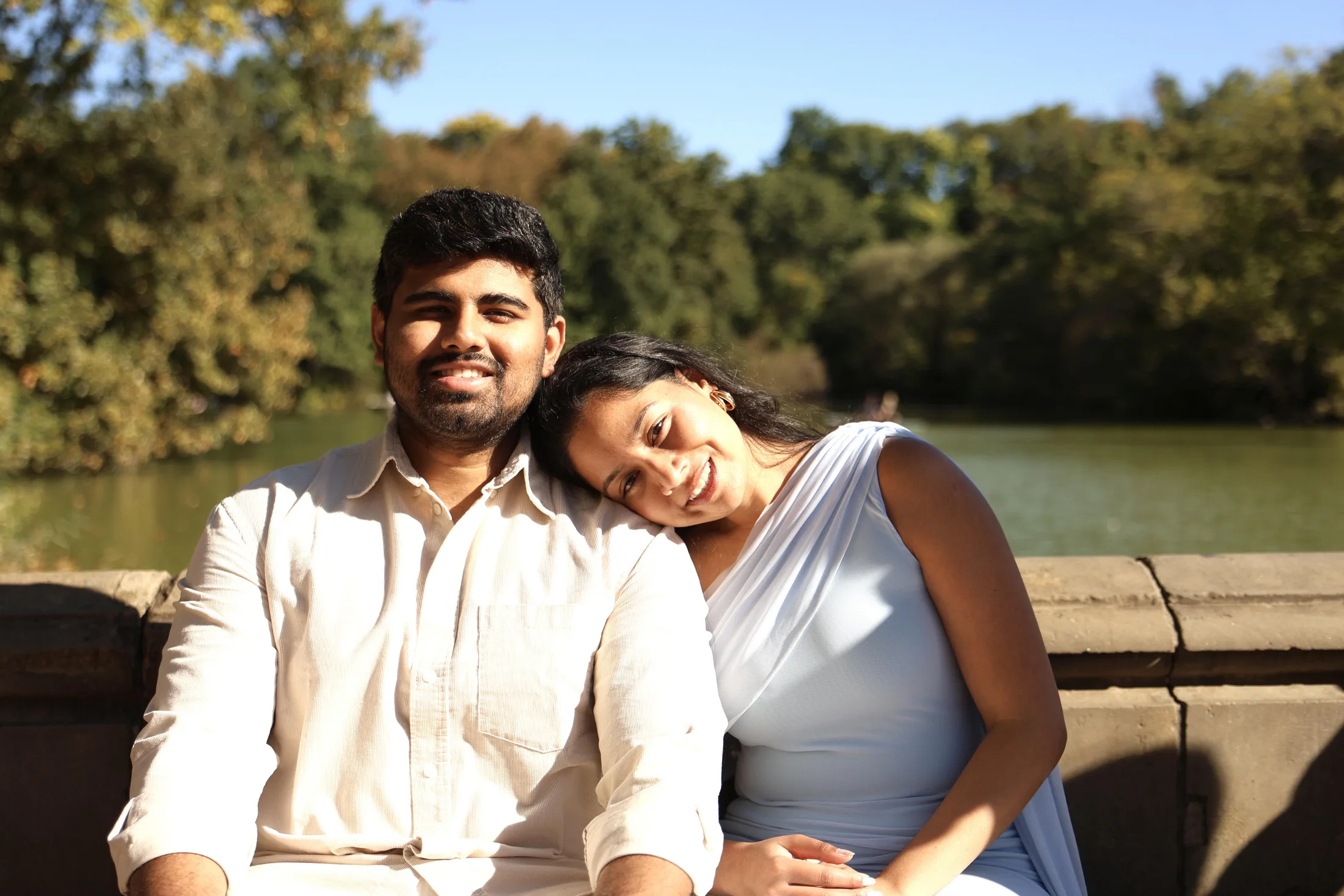 Dakshinya resting her head on Karthik's shoulder as they sit together by the lake in Central Park, both smiling in the bright sunshine