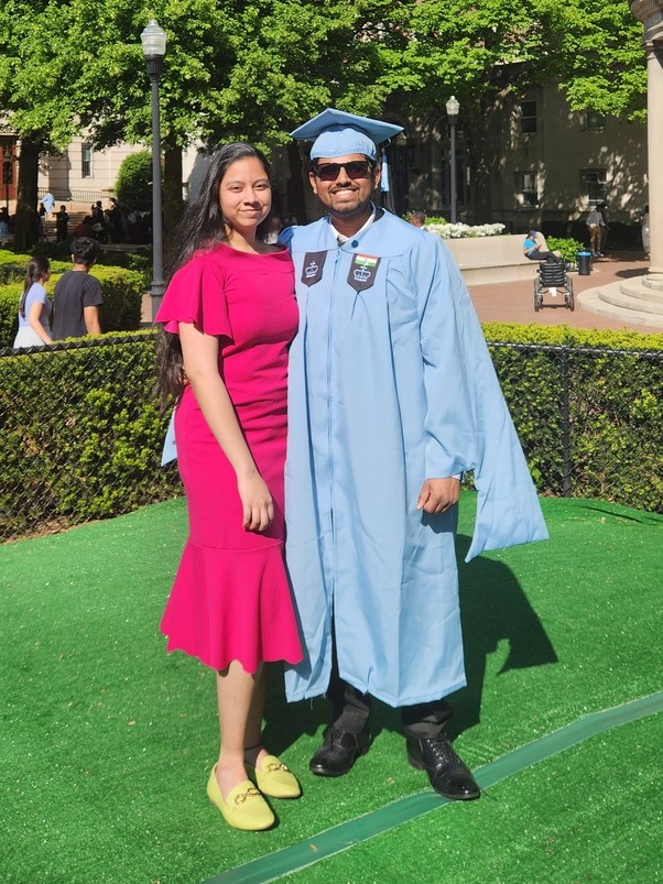 Karthik in a light blue graduation gown and cap standing on the campus lawn with Dakshinya in a pink dress beside him