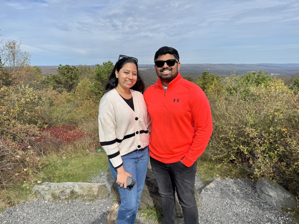 Dakshinya and Karthik standing at a mountain overlook, Karthik in a bright red jacket with rolling hills behind them