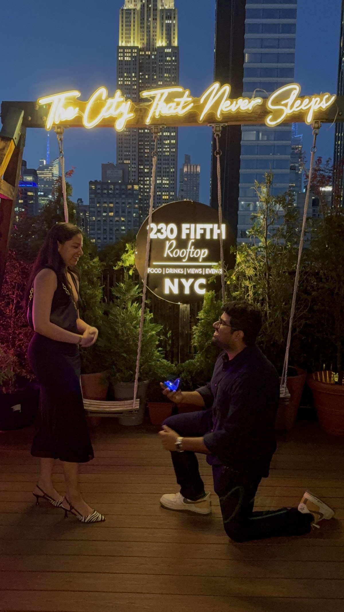 Karthik down on one knee proposing to Dakshinya at a New York rooftop at dusk, the Empire State Building lit up behind them