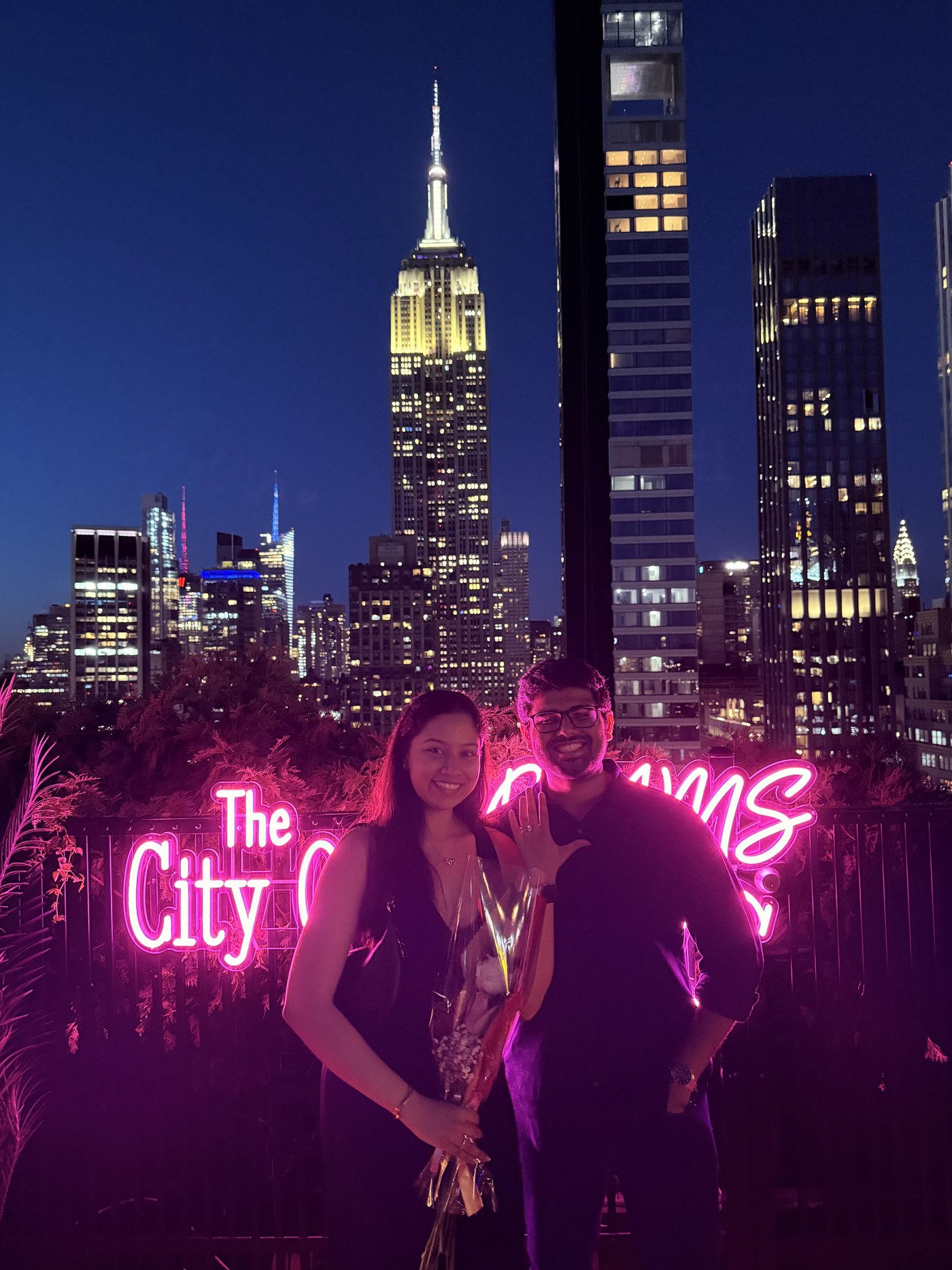 Dakshinya and Karthik smiling in front of a pink neon sign and the New York skyline at night, Dakshinya holding a bouquet and showing her ring